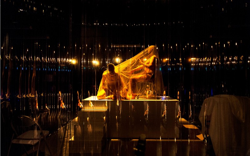 Two performers are situated on a raised platform in a dark room. They are surrounded by mirrors and a golden glow emanates from above them, reflected in each mirror creating an eerie halo of light. One performer kneels on all fours perpendicular to the other, who sits in a chair. They are both wearing a skin tight hood and a flesh coloured leotard. A sheet of transparent tulle is draped over the pair of them.”