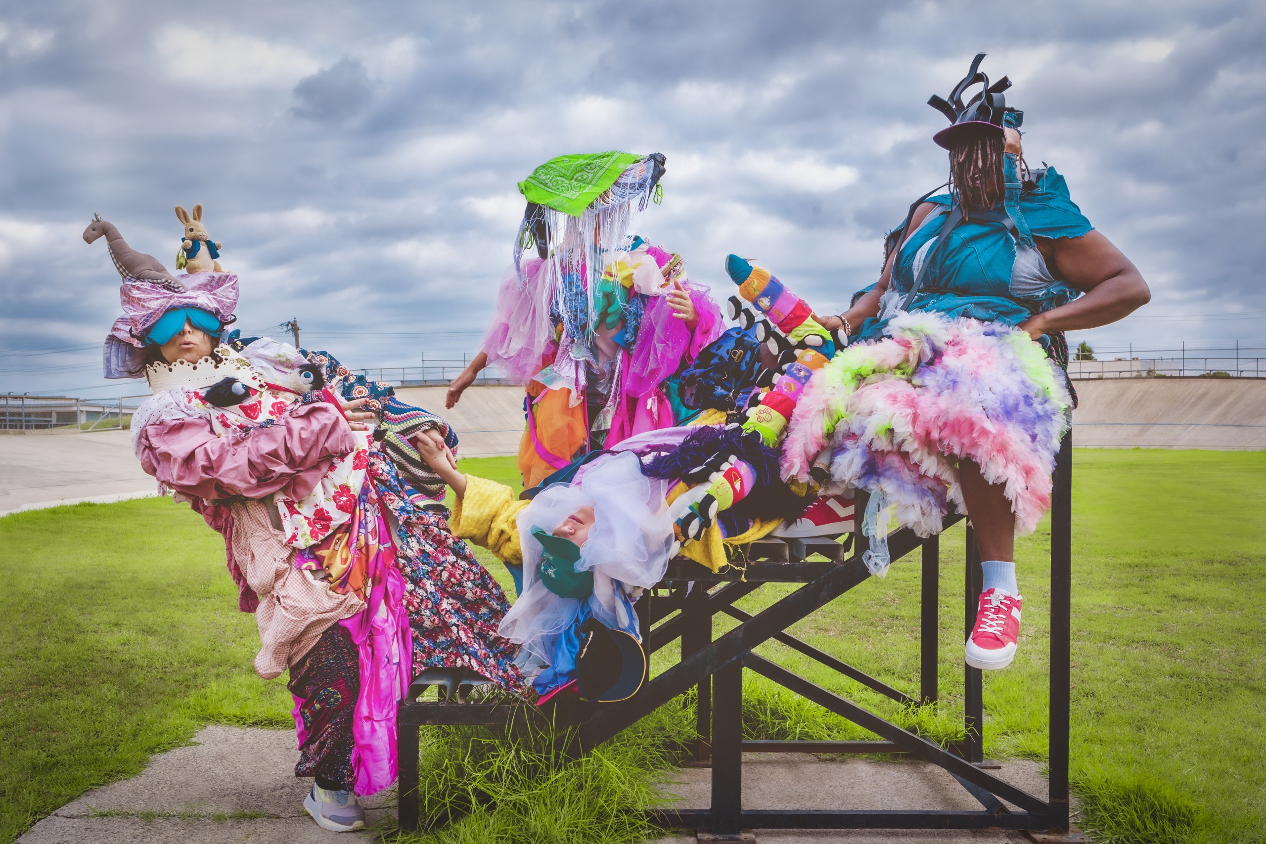 Four performers are draped over an outdoor seating bank in an oval. The sky above them is overcast. Each performer is heavily laden in costumes of layered fabrics, soft toys, tulle etc. Their body shapes are dwarved and faces obscured by the strange costumes and extravagant hats.