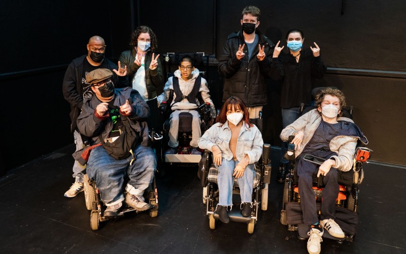 A group shot featuring 17 people who were part of the Artist Lab, posing in front of a black background with five people sitting in wheelchairs at the front.