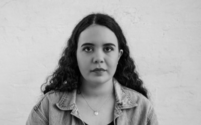 Chloe Ogilvie, a fair skinned First Nations woman, stares direct to camera. She wears a dark t-shirt under a denim jacket. She has long dark hair. The photo is in black and white.