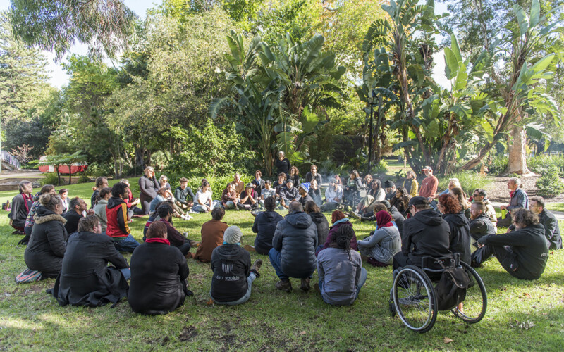 A large and diverse group of artists gather in a circle, seated on the lush green lawn surrounded by nature outside the Subiaco Arts Centre.