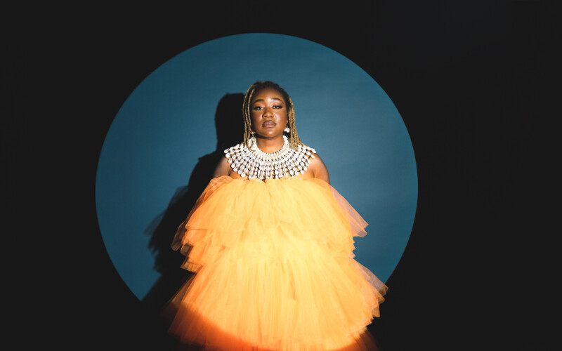 Nancy Denis, a black Haitian-Australian Performer stands in a spotlight staring resolutely to camera. She wears a fabulous full-bodied orange tulle dress and a large ornate necklace made of cowrie shells.
